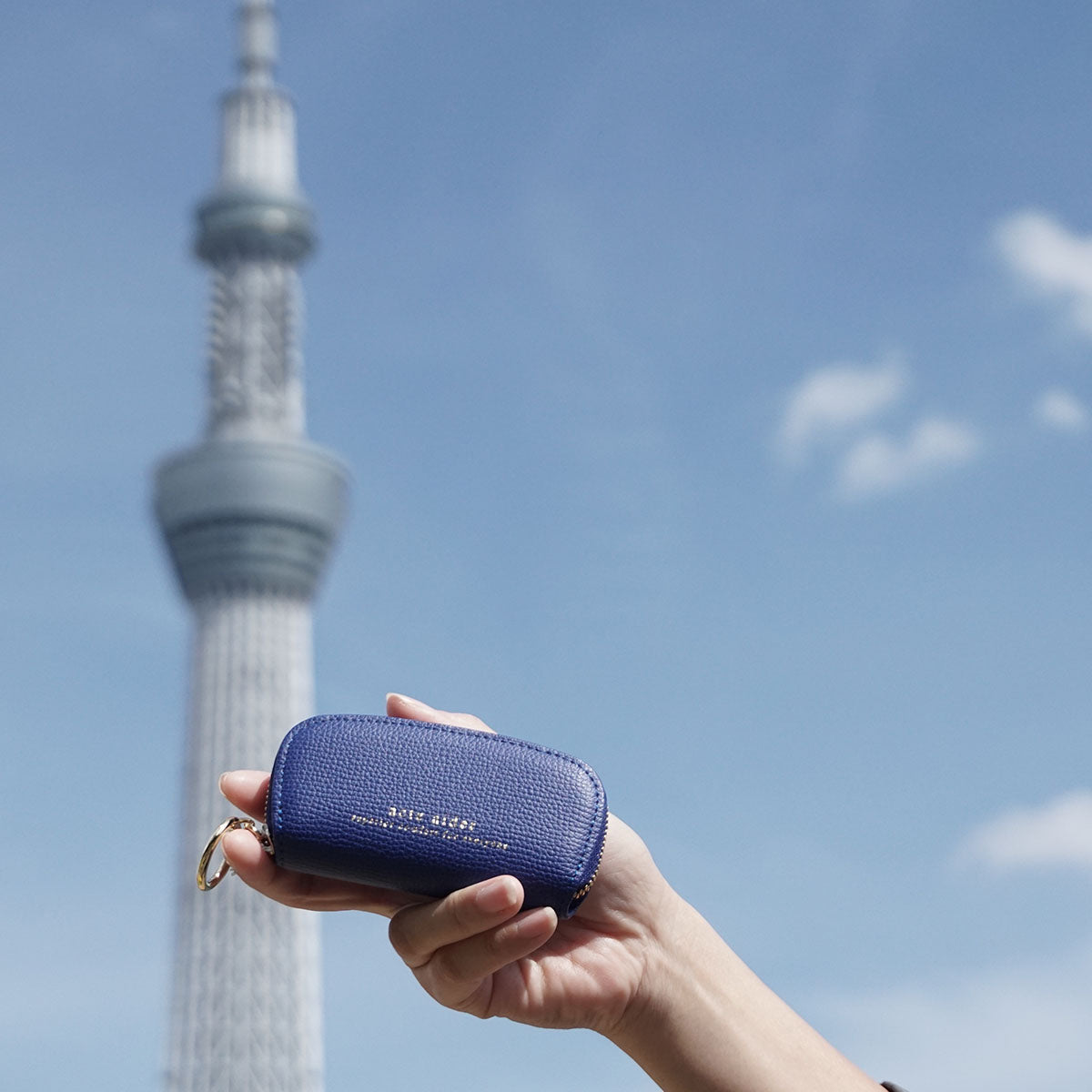 ラウンドファスナー小銭入れ エンボスレザー ブルー アークトエディー 青空とスカイツリーを背景に手にする小銭入れ