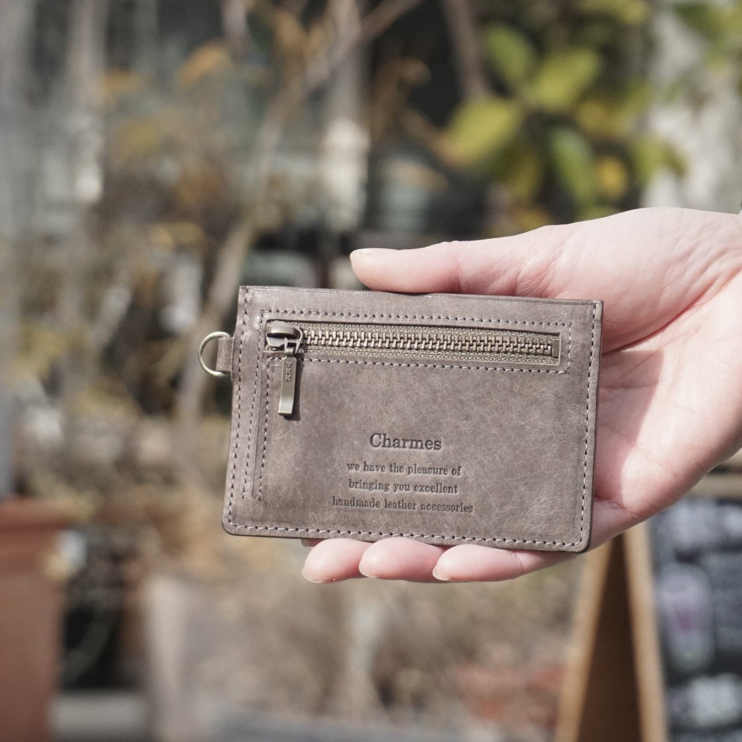 カードケース　マルゴーシリーズ　グレー　シャルム　駒屋　陽射しの当たるカフェの前で右手にカードケースを持ち、こちらに見せている手元のアップ画像