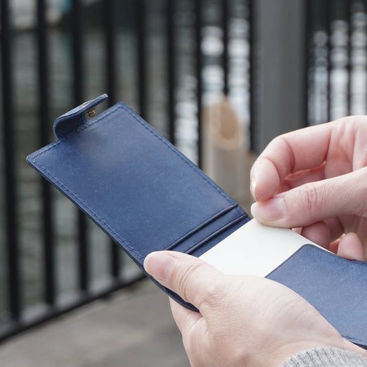 縦型カードケース　マルゴーシリーズ　ブルー　シャルム　駒屋　川のほとりでカードケースを開き、そこから1枚のカードを取り出そうとしている様子　手元のアップ画像